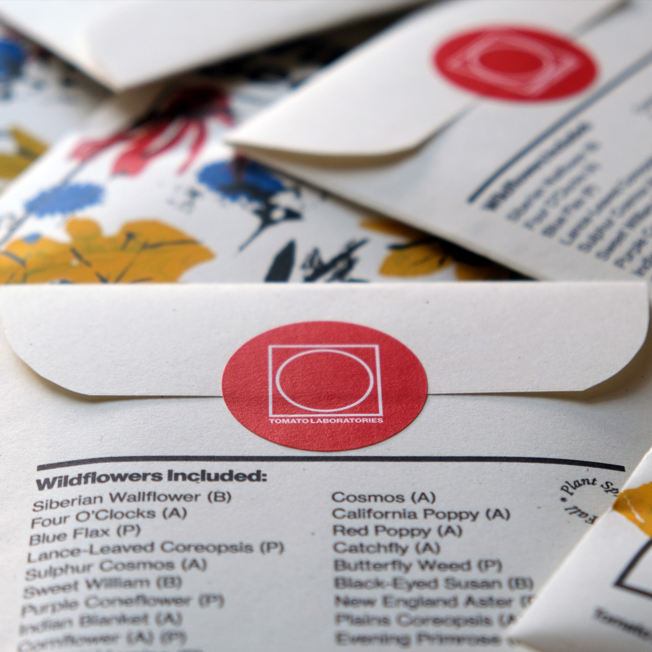 A loose assortment of wildflower seed packs is scattered and in focus is a bright red sticker on the back of a pack with the tomato lab logo on it