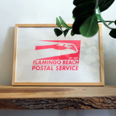 A neon pink flamingo beach postal service print sits on white paper in a wood frame on a wooden bench with a plant over it