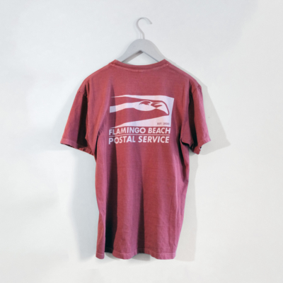 A flamingo colored pocked tee shows a large Flamingo Beach Postal Service logo in white on the back as it hangs on a white wall