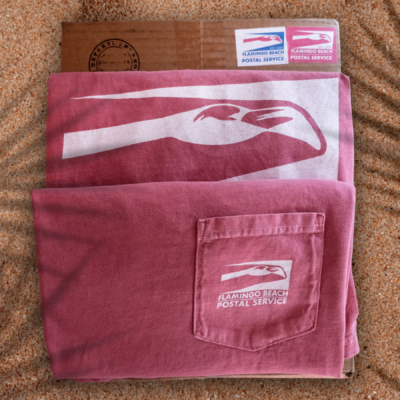 A flamingo pink, folded pocket tee with a small white Flamingo Beach Postal Service logo over the breast pocket sits neatly folded on a cardboard box with stamps on stop of a sandy boardwalk under some palm frond shadows