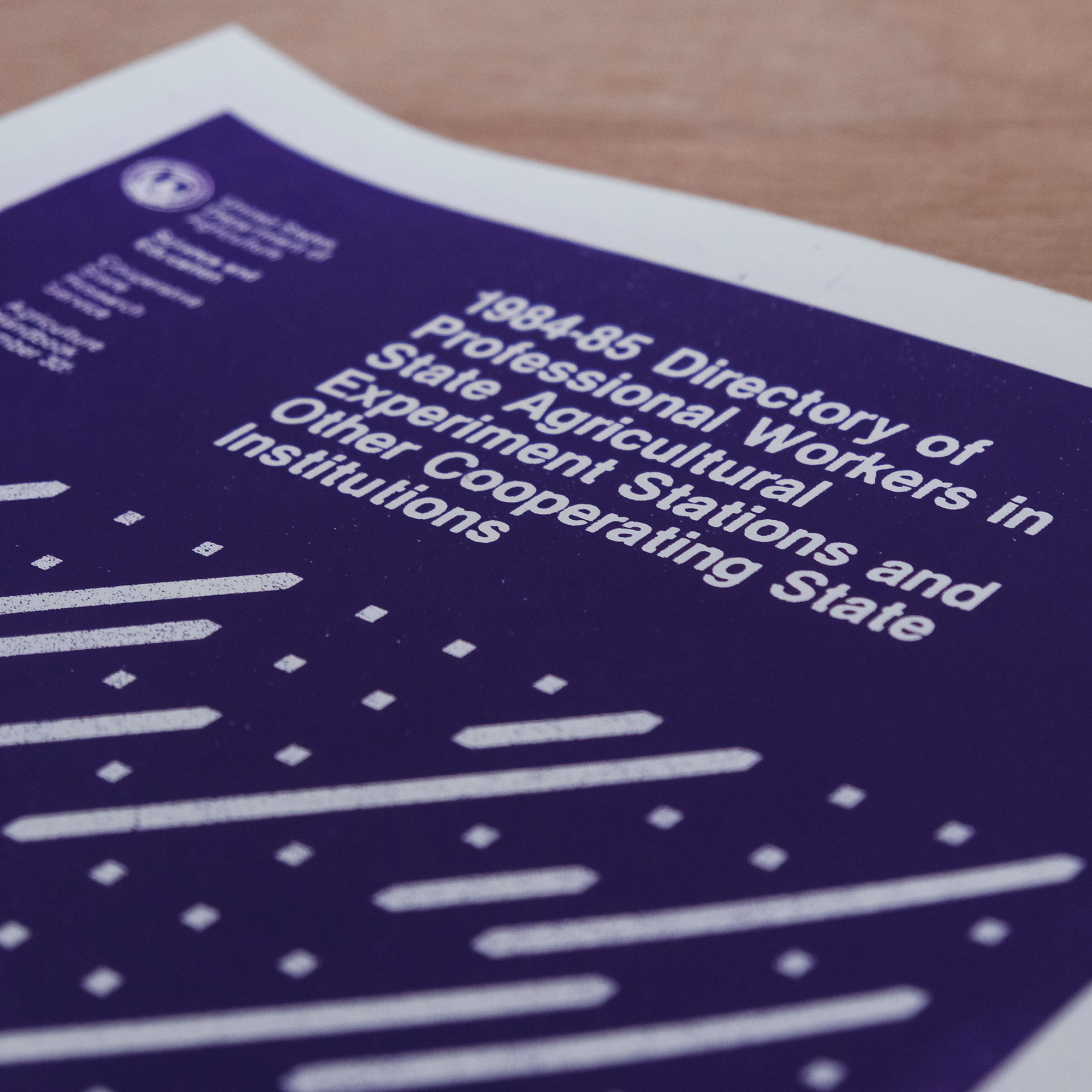 A purple silkscreened print of USDA report 1984-85 Directory of Professional Workers in State Agricultural Experiment Stations and Other Cooperating State Institutions which has geometric shapes in white and purple and the print sits on a wood surface