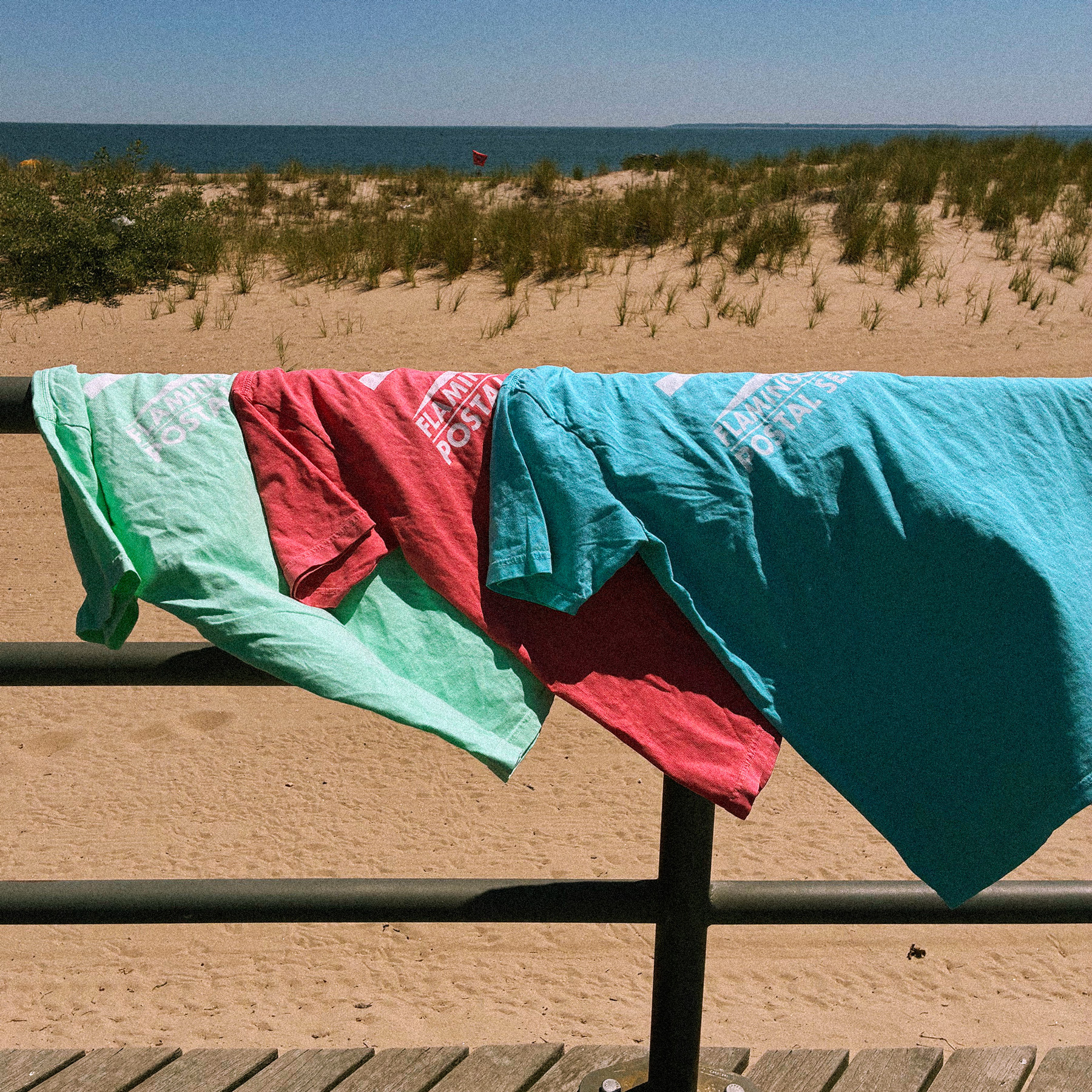 A flock of Flamingo Beach Postal Service tees drapes lazily over a beach railing out down by the boardwalk, colors glowing and radiating into the sunny beach air