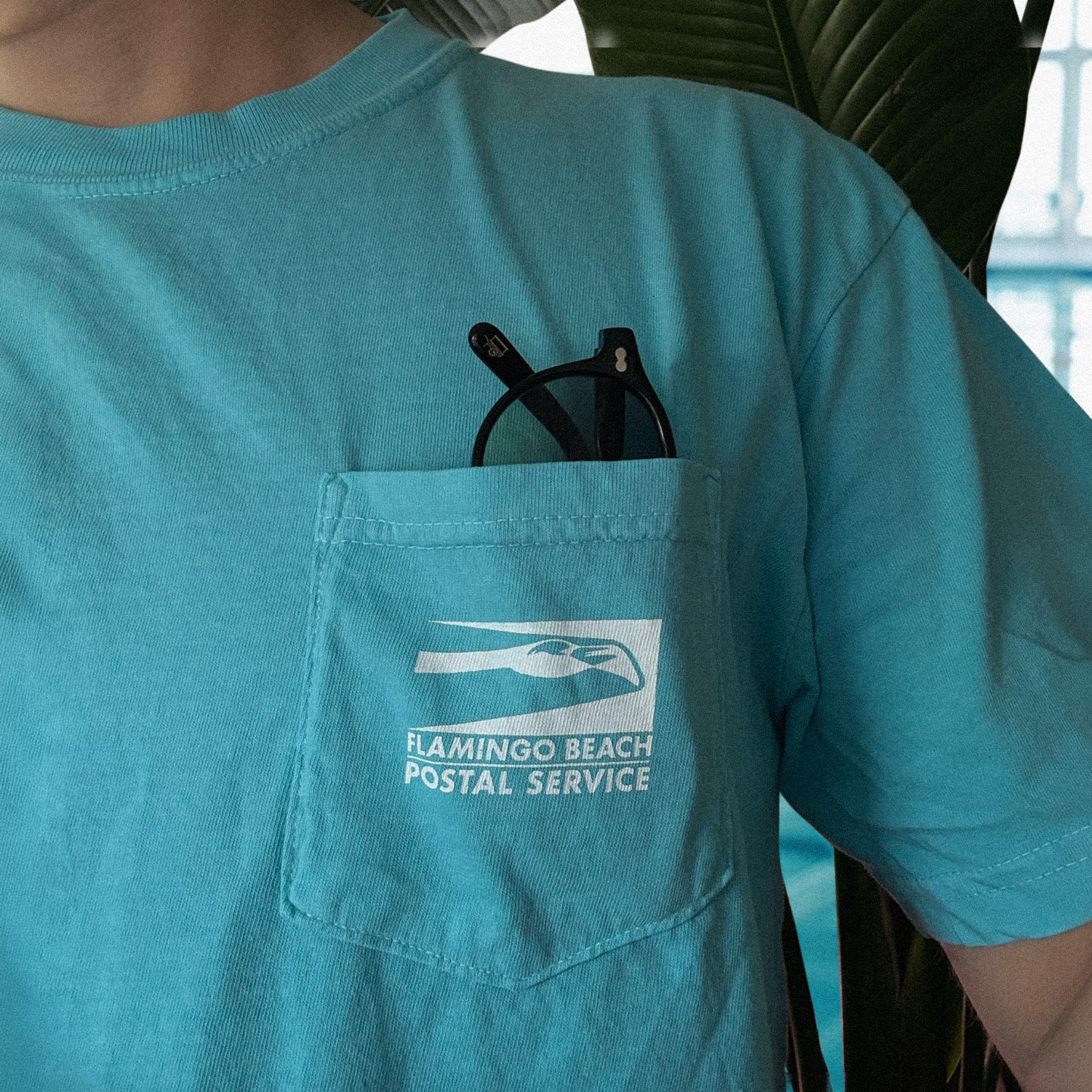 A man in a pool house wearing a Lagoon colored pocket tee with the Flamingo Beach Postal Service logo in white on the pocket