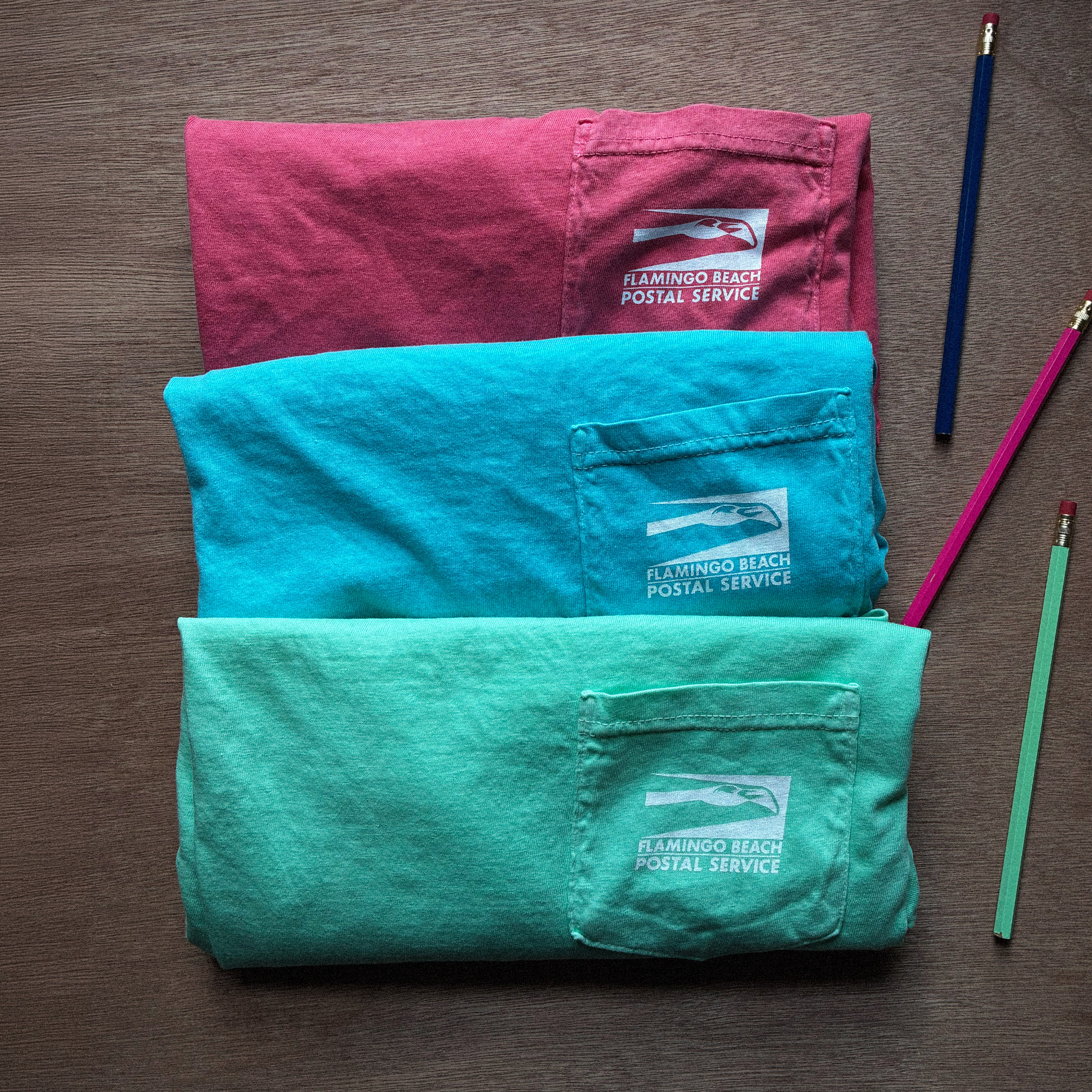 A trio of colorful pocket tees featuring the Flamingo Beach Postal Service logo