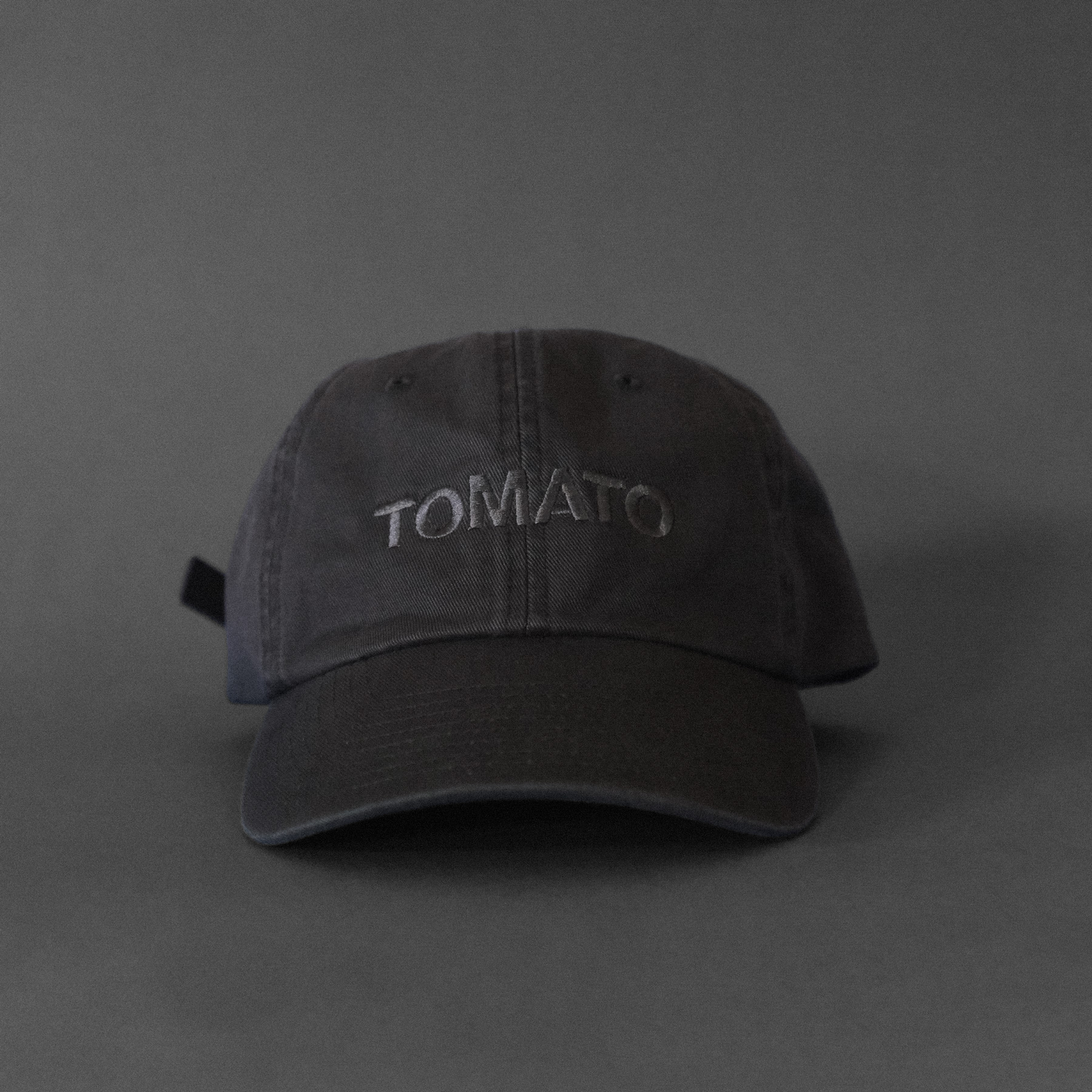 A dark gray cap with dark gray embroidered text that says TOMATO on a dark gray background