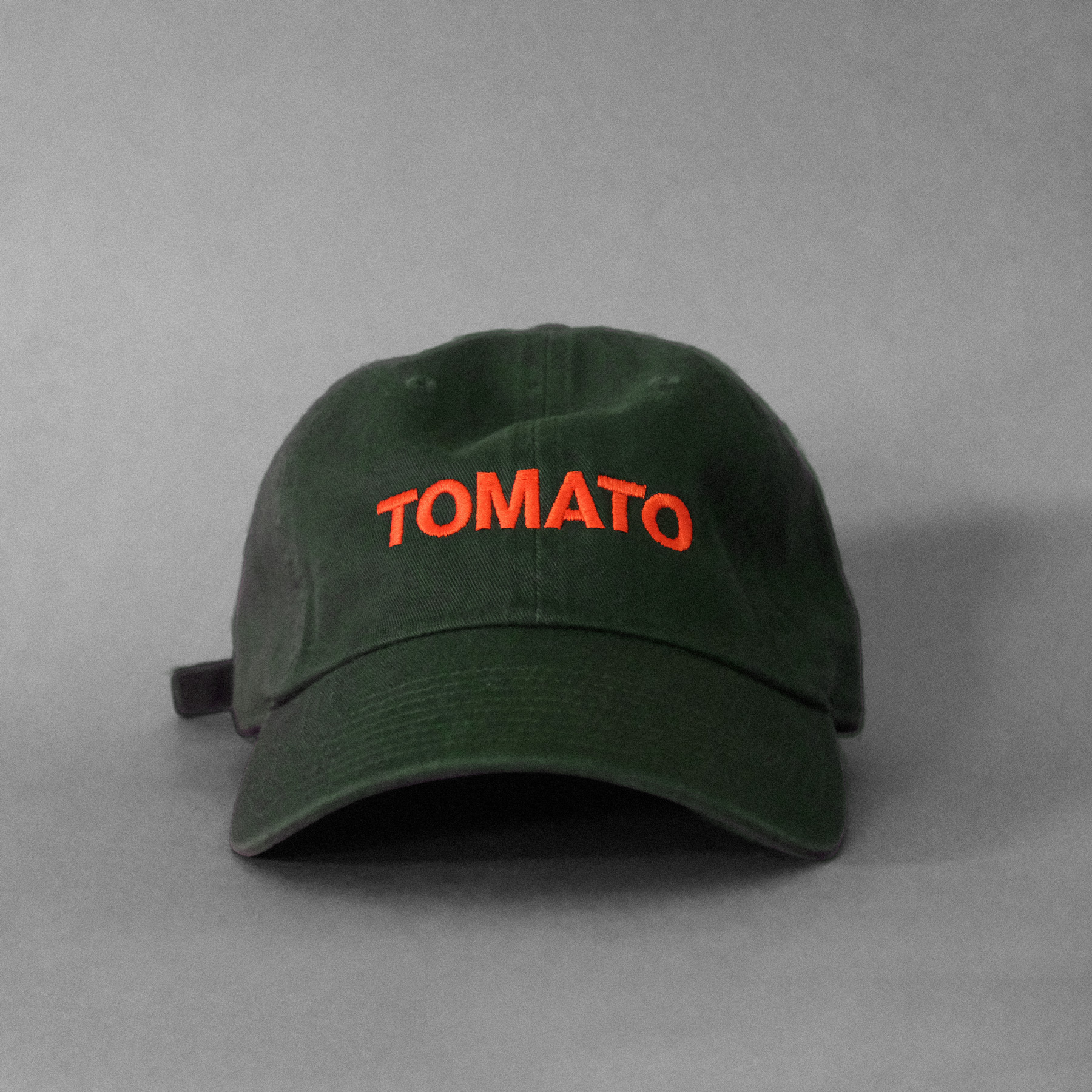 An olive cap with orange embroidered text that says TOMATO on an gray background