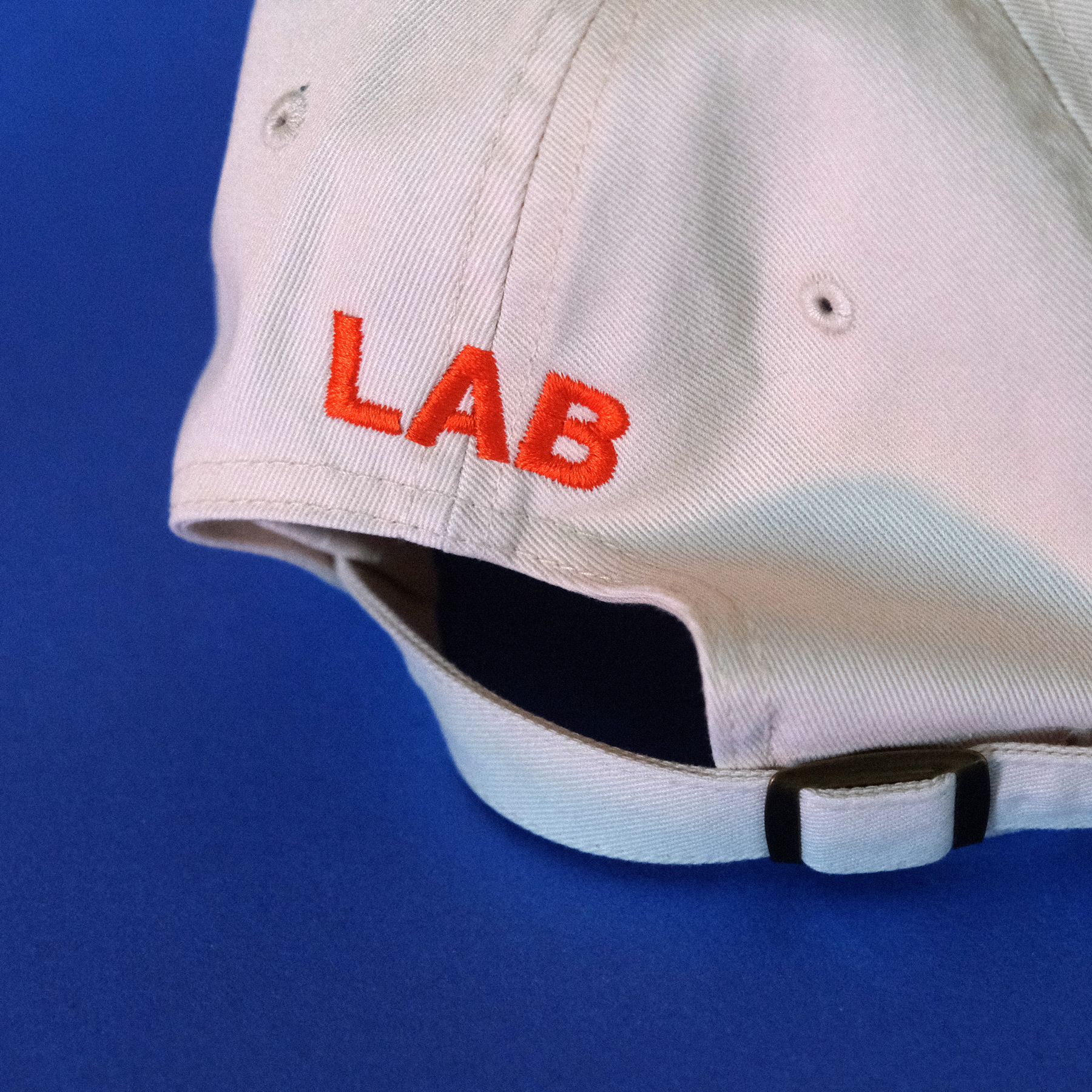A natural light cap with orange embroidered text that says LAB on a blue background