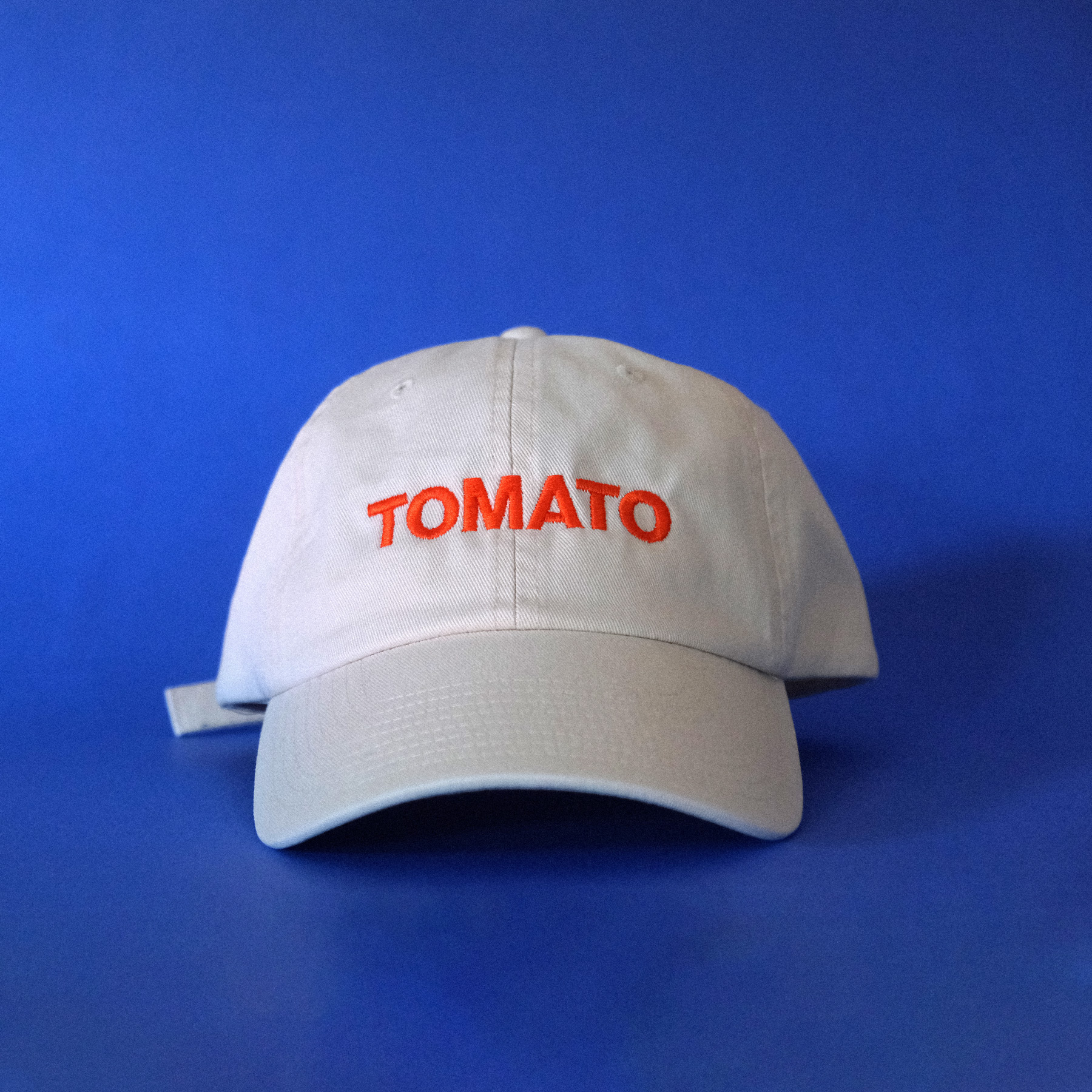A natural light cap with orange embroidered text that says TOMATO on a blue background